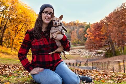 A smiling woman wearing eye glasses and a hat sitting on the ground holding a dog with fall foliage trees in the background.
